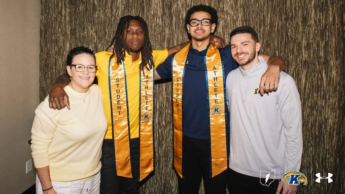 Jamal Sumlin and Brenden Moss of Kent State Men's Basketball wearing yellow stoles with "Student Athlete" printed on them. Academic Counselor Jana Ashley and Assistant Coach Tommy Luce are on each side of them. They are indoors against a textured wall. The Under Armour and Kent State University logos are visible in the bottom right corner.