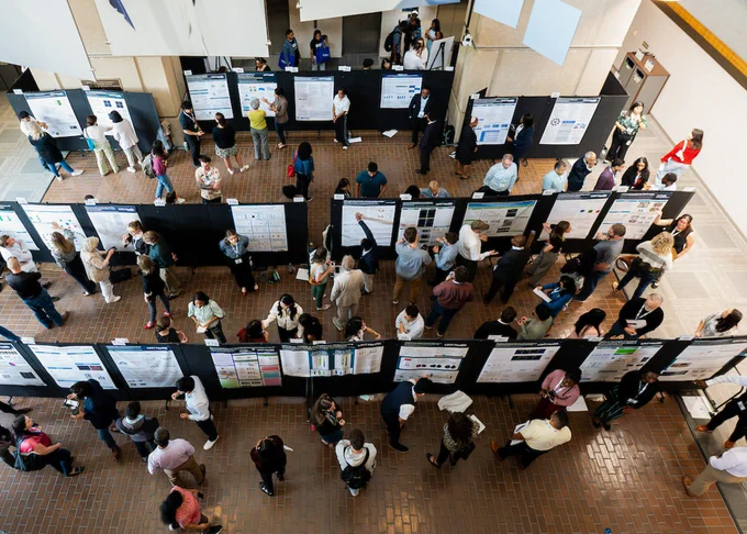 Aerial view of a bustling conference hall with numerous people examining research posters displayed on large boards. The scene captures a lively academic atmosphere with attendees engaged in discussions and networking.
