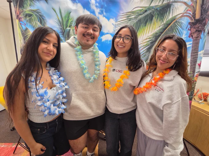 A group of four people wearing leis and casual clothing stand smiling in front of a backdrop featuring palm trees and a blue sky with clouds.