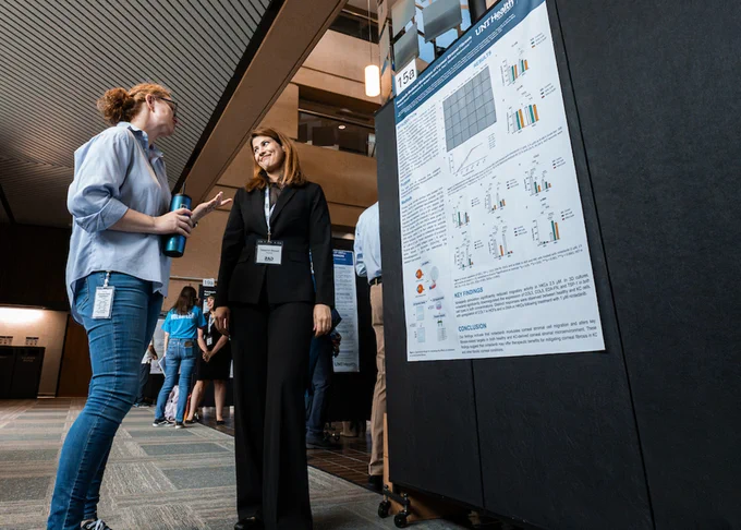 Two individuals stand in front of a scientific poster display. The poster includes graphs and text. They appear to be in a conference or professional setting with others in the background.