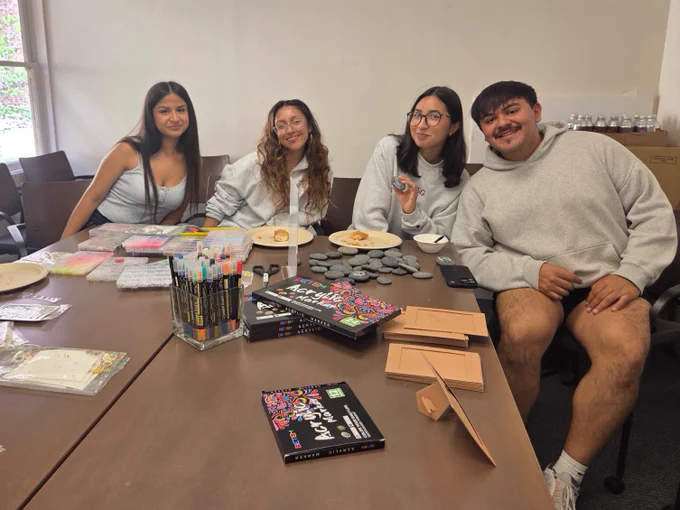 A group of four people sitting at a table filled with art supplies, books titled "The Art of Zines," and decorated stones. They are smiling and appear to be in a creative workshop or art session.