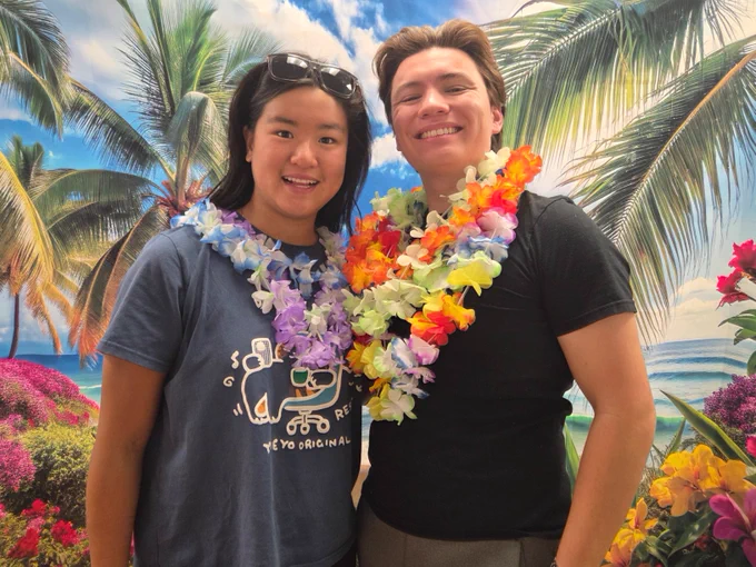 Two people stand smiling in front of a tropical background with palm trees and ocean waves. They are wearing colorful leis. The individual on the left is wearing a shirt with a graphic. The scene is vibrant and bright, suggesting a relaxed, sunny atmosphere.