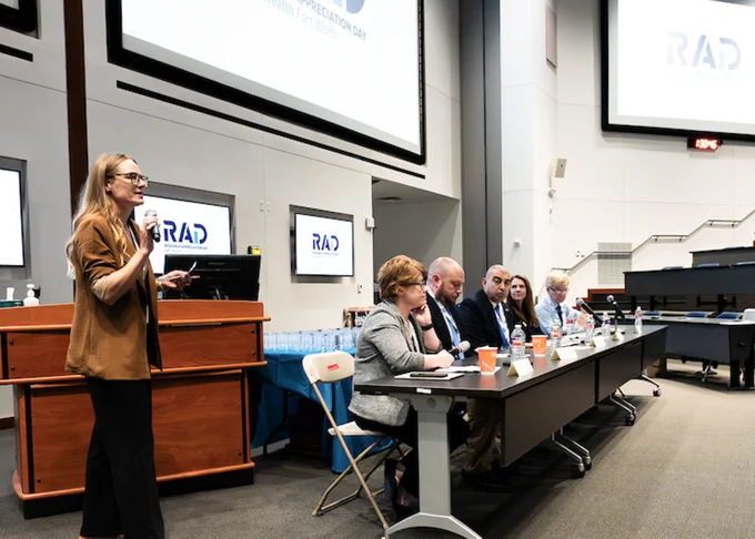 In a conference room, a person is standing and speaking into a microphone at a podium. Five people are seated at a long table with microphones and laptops, engaged in a panel discussion. Multiple screens display the word "RAD."