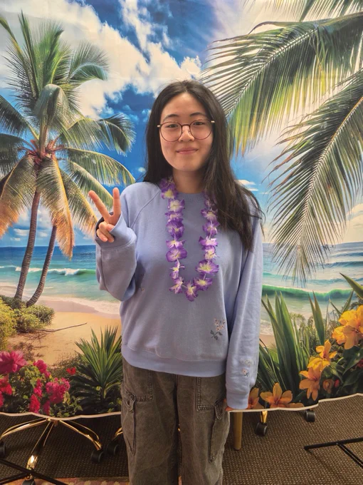 A person stands in front of a colorful beach-themed backdrop featuring palm trees, a sandy shore, and blue skies. They are wearing a light blue sweater, gray pants, and a purple flower lei, while making a peace sign with one hand.