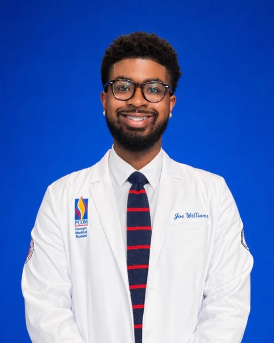 A person smiling at the camera sitting against a blue background. They are wearing a white coat and a striped tie.