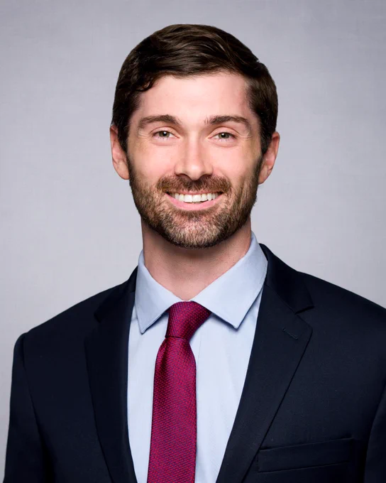 A person smiling at the camera sitting against a white background. They are dressed professionally, wearing a red tie and black jacket.