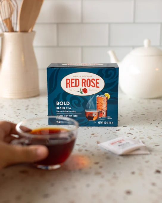 Box of Red Rose Bold Black Tea on a kitchen counter with a hand holding a cup of brewed tea in the foreground.