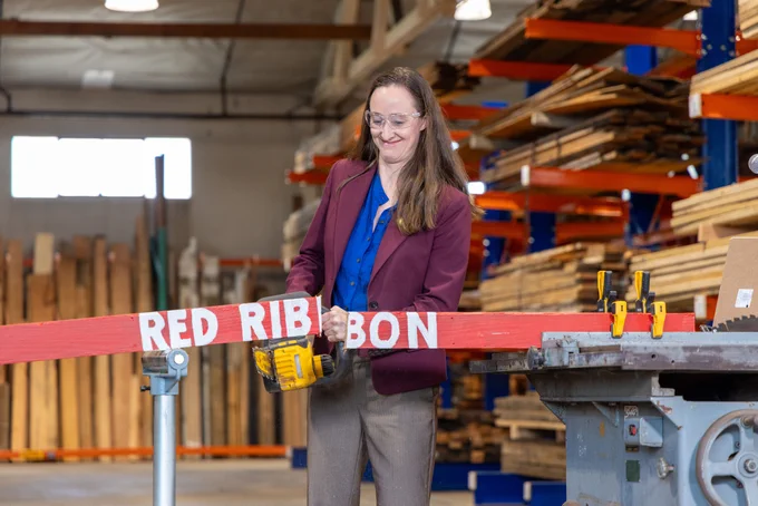 A person is using a power tool to cut a red ribbon labeled "RED RIBBON" in a workshop with stacked lumber and shelving in the background.
