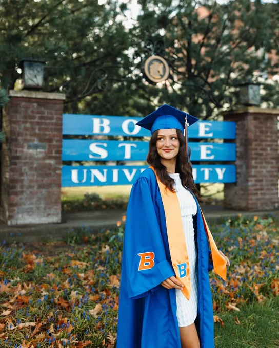 Boise State student smiling wearing their blue cap and gown in front of the Boise State sign for their graduation photos. 
