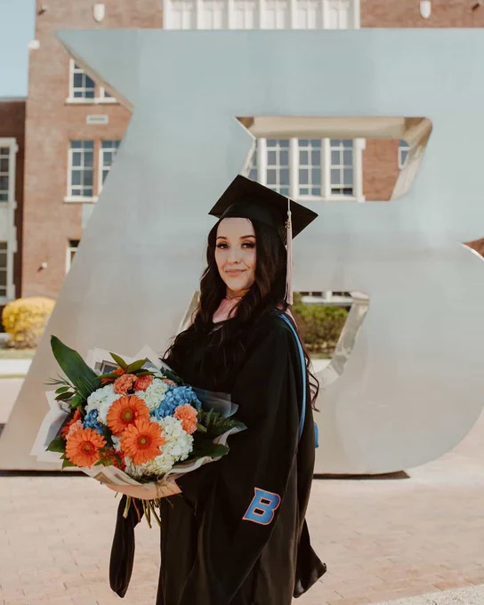 Boise State graduate student holding a blue and orange bouquet of flowers wearing masters degree robes in front of the B Plaza. 

