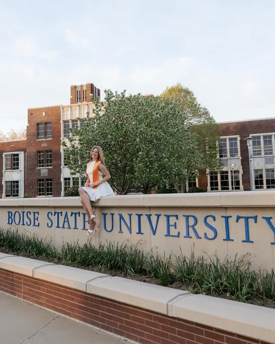Boise State student sitting and smiling on the Boise State University sign at the B plaza. 
