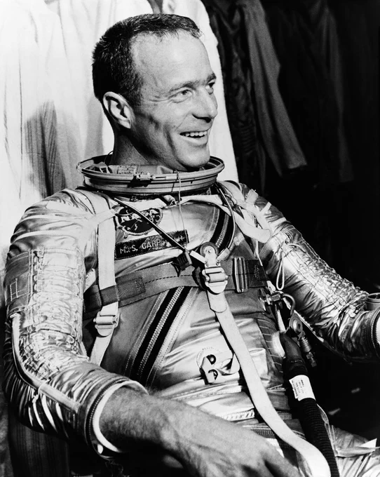 Black and white image of a man in a shiny spacesuit without  a helmet. He is smiling and looking off-camera.
