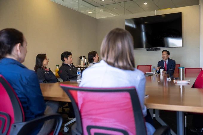 A group of people engaged in a meeting around a large conference table. A person at the head of the table appears to be speaking. A large monitor is mounted on the wall in the background. The setting suggests a professional environment.