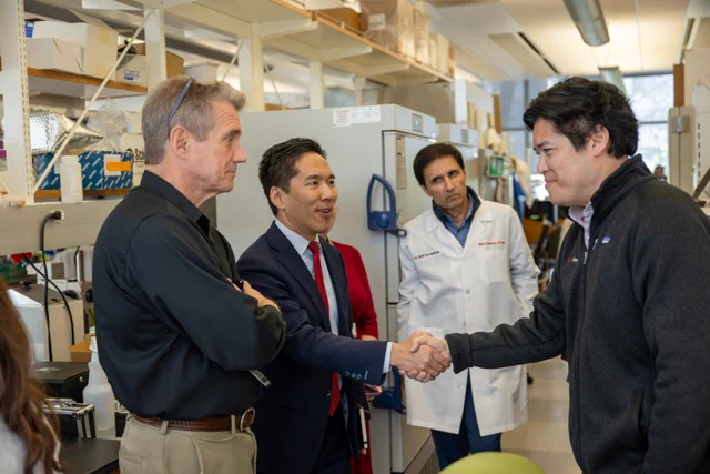 A group of people are in a laboratory setting. One person is wearing a lab coat, and two others are in business attire, shaking hands with another individual in a casual jacket. Lab equipment and shelves are visible in the background.