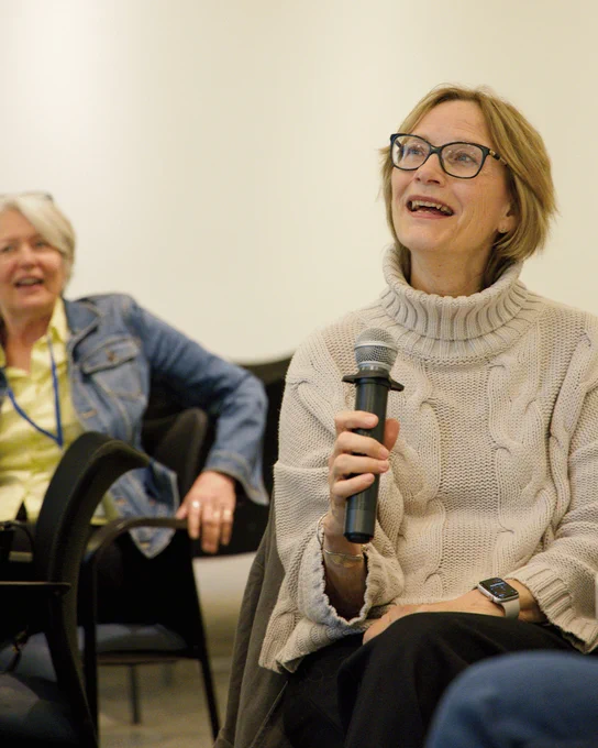 A smiling woman with chin-length blonde hair wearing glasses and a knit sweater speaks into a handheld microphone