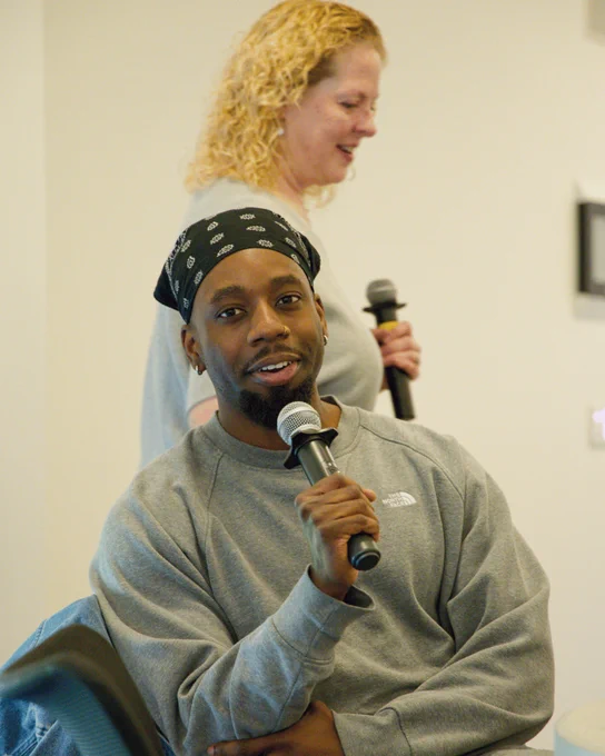 A smiling man with a black chin beard and wearing a gray sweatshirt and a black bandana on his head speaks into a handheld microphone