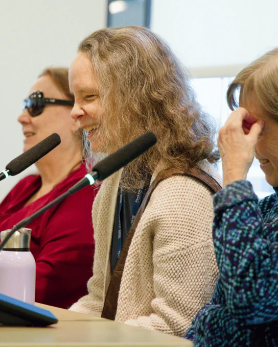 Three women sitting at a table with microphones on it laugh 