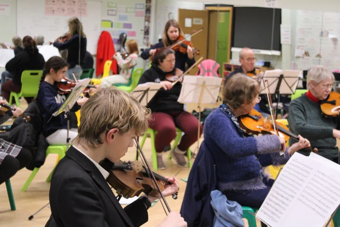 A group of musicians seated in a classroom setting, playing violins and reading sheet music during a rehearsal.