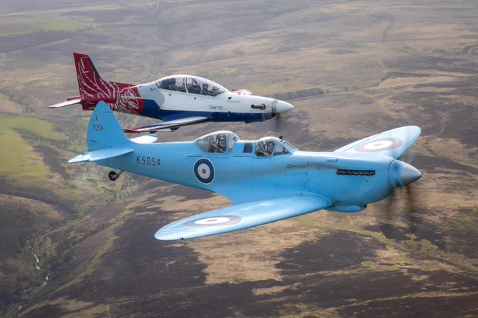 A blue Spitfire flies alongside a red and white Qinetiq aircraft.