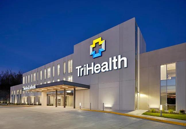 Exterior view of a modern TriHealth medical office building at dusk, with a large “TriHealth” sign and logo on the facade. The building features clean lines, large windows, a lit entrance canopy, and a paved driveway in the foreground.