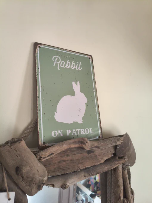 A green metal sign with a silhouette of a white rabbit sitting in the middle. Above and below, text reads, Rabbit on patrol. It is stood on top of a driftwood mirror.