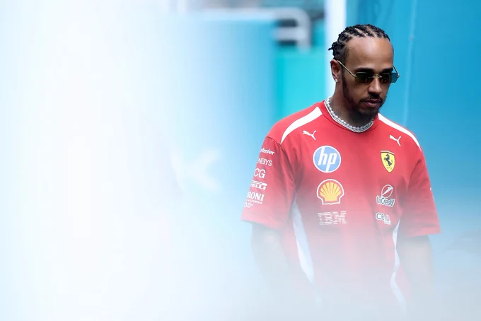 A photo of Lewis Hamilton looking towards the ground as he walks. He is wearing sunglasses and a red shirt with various logos on it.