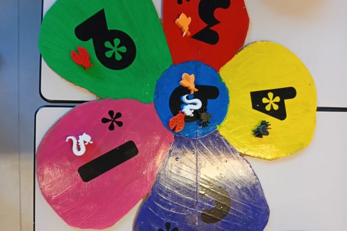 Plateau de jeu coloré en forme de fleur, fabriqué par des élèves, avec des pétales numérotés et décorés de petits insectes et animaux, posé sur une table en classe.