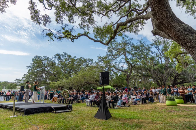 A person stands on an outdoor stage under large trees, addressing a seated audience.