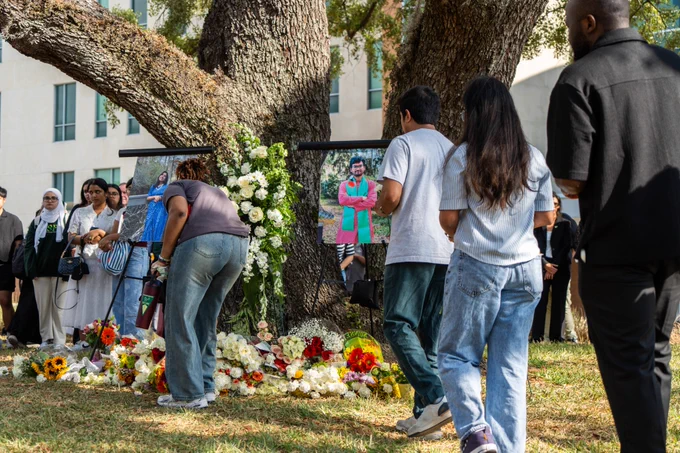 A group of people gather around a memorial beneath a large tree, featuring flowers and photos.
