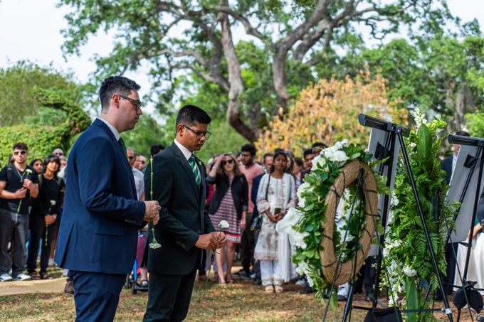 A group of people is gathered outdoors for a memorial or ceremony. Two individuals in formal attire stand at the forefront, holding flowers, near a display of wreaths and floral arrangements. A larger crowd is visible in the background, surrounded by greenery and trees.