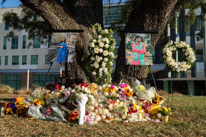 A memorial site under a large tree features two framed photos on easels. The area is adorned with numerous bouquets of colorful flowers, a white floral wreath, and two lit candles.
