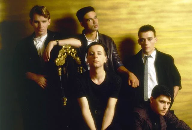 Simple Minds posed in a studio promo shot around 1982, featuring five band members against a warm golden backdrop. Jim Kerr sits front and centre with an intense, focused expression, while the rest of the band stand and lean around him in sharp, dark attire, capturing the confident, rising energy of their New Gold Dream era.