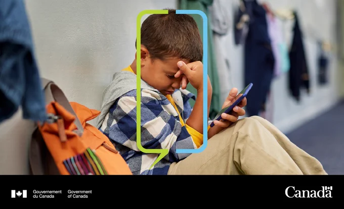 ALT : Un jeune enfant assis par terre dans un couloir d’école, son sac à dos à côté, regarde son téléphone, l’air en détresse. 

