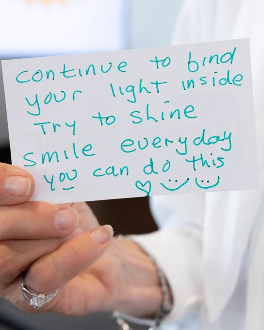 A person is holding a handwritten note that reads, "Continue to find your light inside. Try to shine, smile every day. You can do this." The note includes a heart symbol and two smiley faces. The background is indoors and slightly blurred.