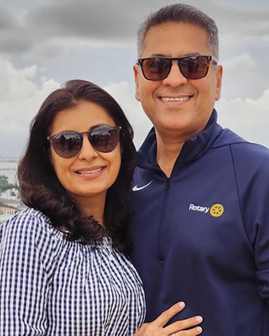 An image of two people posing outdoors, both wearing sunglasses. One person wears a blue shirt with the Rotary International logo. 