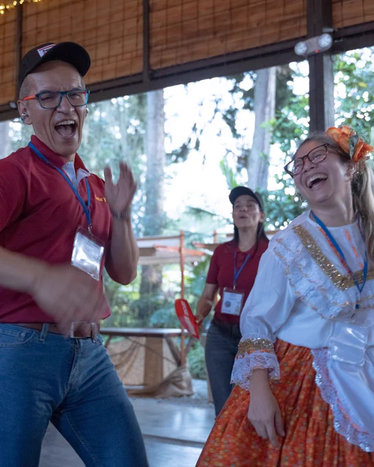 A group of people joyfully dancing indoors. One person wears a red shirt and a black cap, smiling, while another wears a colorful traditional outfit with orange accents and floral decorations in the hair. The background is airy with large windows showing greenery outside.