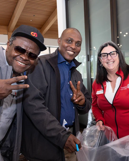 Three individuals are smiling while participating in a community service activity indoors. Two are gesturing with peace signs. One person is wearing a badge with a Rotary Club logo. The setting appears to be a modern building with large windows and wood accents.