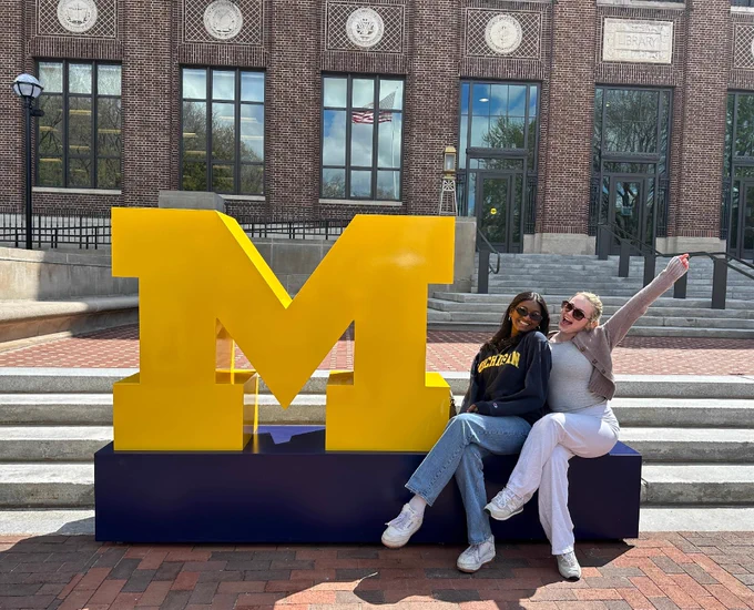 Two friends sitting next to a giant block M in front of the U-M Hatcher Library.