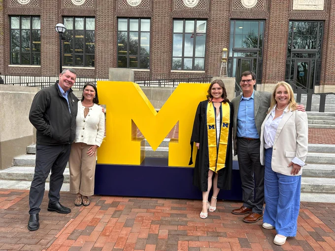 A family standing on either side of a giant block M in front of the U-M Hatcher Library.