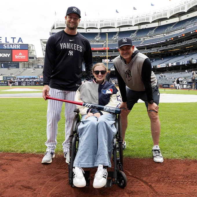 Gerrit Cole and Aaron Boone present Sarah Langs with an autographed Lou Gehrig Day baseball bat on the field at Yankee Stadium. Cole wears a black Yankees long sleeve shirt, pinstriped pants and a dark blue Yankees cap. Boone wears a black long sleeve shirt, gray hooded Yankees sweatshirt tank, dark gray shorts and a dark blue Yankees cap. Langs wears denim jeans and an MLB jacket with sunglasses. The bat is red and dark blue with a Lou Gehrig Day logo on it.