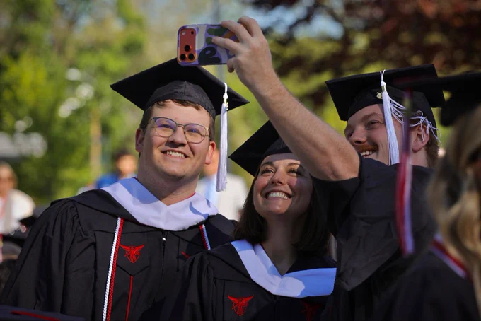 Three graduates in black caps and gowns smile while taking a selfie outdoors at a commencement ceremony, with green trees visible in the background.