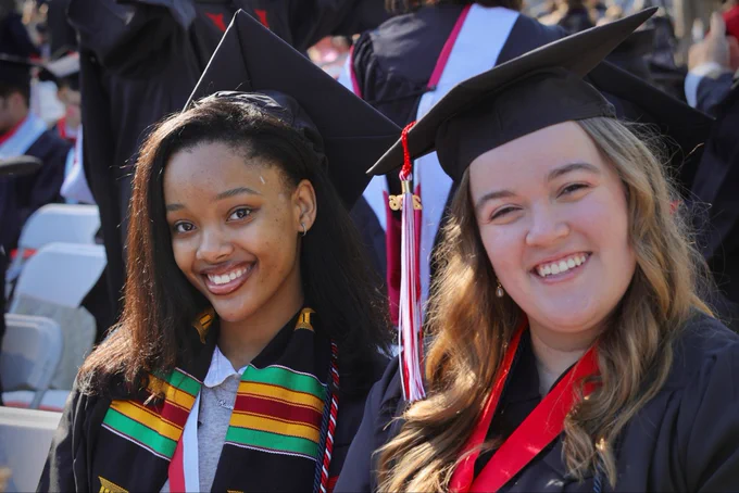Two smiling graduates in black caps and gowns pose together at an outdoor commencement ceremony, one wearing a colorful kente stole and the other wearing a red ribbon.