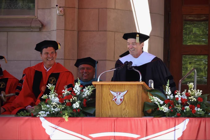 Hugh Jackman in academic regalia addresses the audience from a flower-adorned podium bearing the Ball State University logo during an outdoor commencement ceremony, while university officials seated nearby smile and laugh.