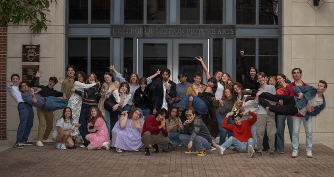 A diverse group of people enthusiastically posing for a photo outside a building marked "College of Motion Picture Arts." They display various energetic and playful poses, with some sitting on the ground and others being lifted off the ground. The setting is casual and joyful.