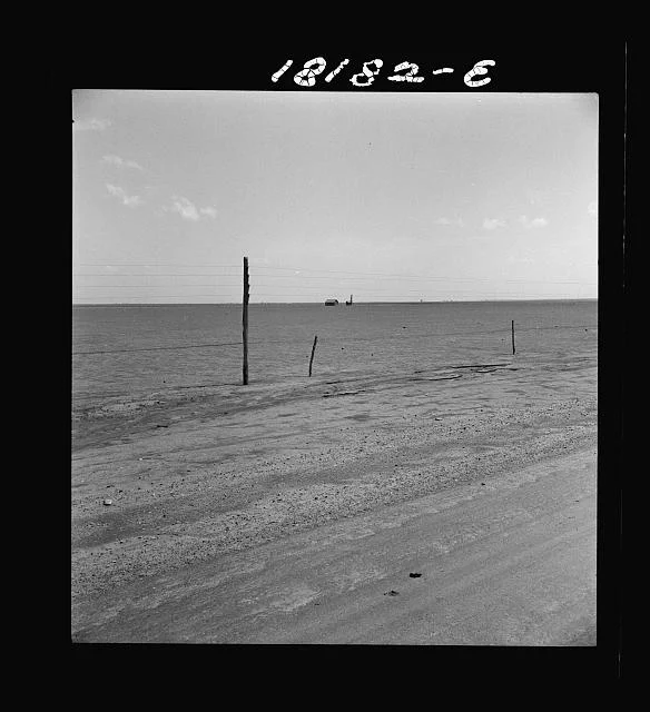 Farm Security Administration photograph of an abandoned farm in Texas County, Oklahoma. An abandoned wood-frame house sits in a dust field behind a barbed wire fence. A powerline is hanging along the top of the scene.