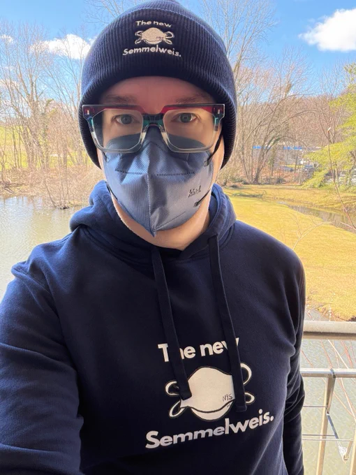 Photo of wire man wearing a dark navy beanie with drawing of a white n95 respirator with the words the new Semmelweis in white text surrounding it. The man is wearing red green and clear glasses with a dark blue kn95 respirator and dark navy drawstring hoodie with drawing of a white n95 respirator with the words the new Semmelweis in white text surrounding it. Background of water, trees and blue and cloudy sky is blurred.