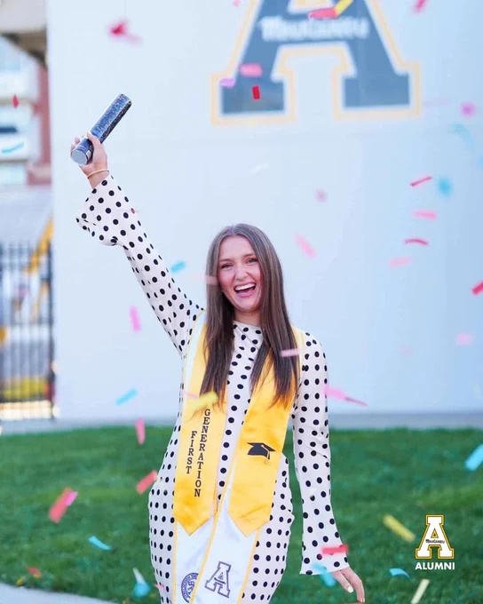 Brittany Senior celebrates in front of a large App State “A” while holding a confetti popper, wearing a yellow first-generation stole and white dress with black polka dots.