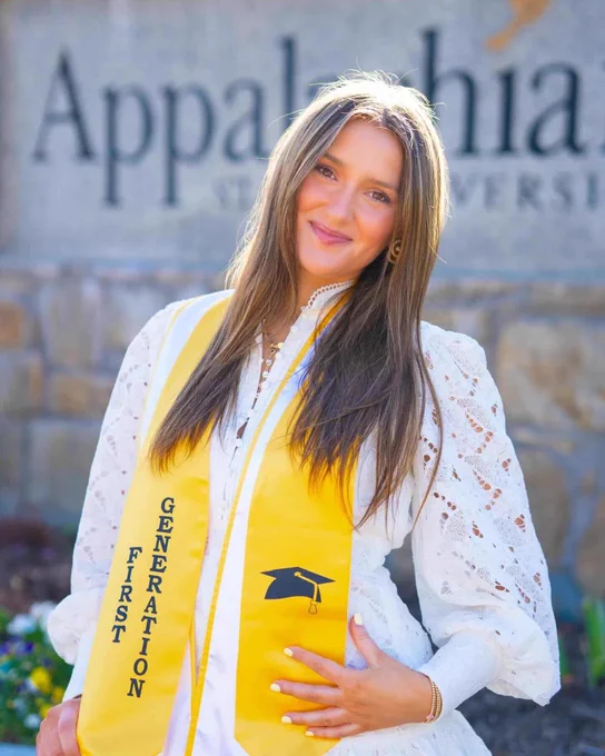 Brittany Senior smiles in front of the Appalachian State University sign, wearing a white dress and yellow first-generation stole.
