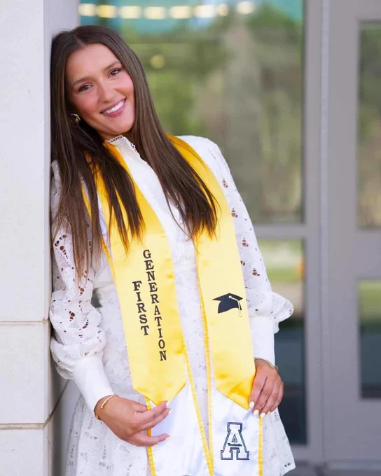 Brittany Senior leans against a campus building doorway, smiling while wearing a white dress and yellow first-generation stole.
