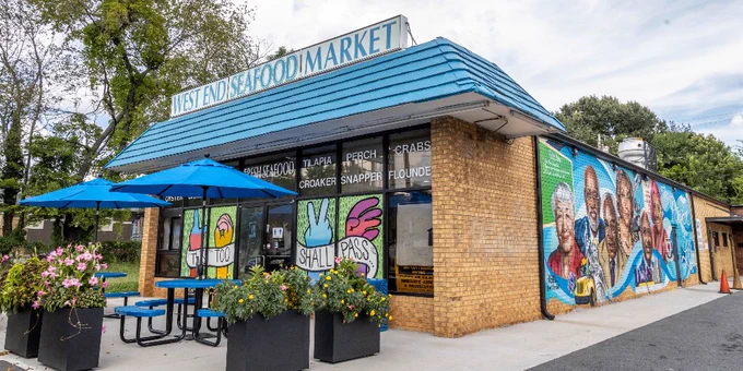 This image shows the West End Seafood Market, a brick building with a blue roof. The storefront features a vibrant mural depicting diverse historical figures. A picnic table with blue umbrellas and planters is in front.